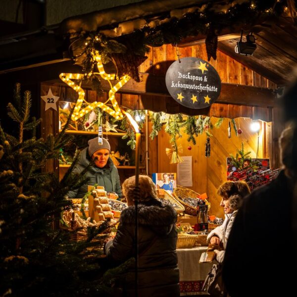 Die Menschen stehen an einem warm beleuchteten, mit Sternen und Grünzeug geschmückten Holzmarktstand und kaufen abends bei einem Verkäufer Backwaren.
