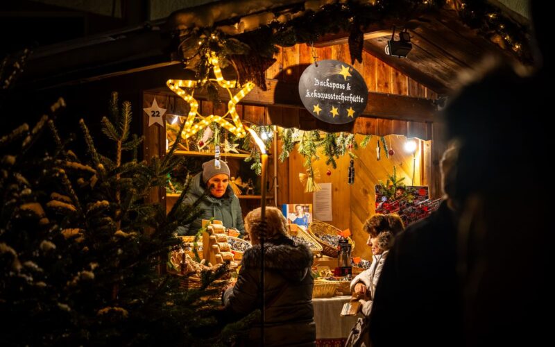 Die Menschen stehen an einem warm beleuchteten, mit Sternen und Grünzeug geschmückten Holzmarktstand und kaufen abends bei einem Verkäufer Backwaren.
