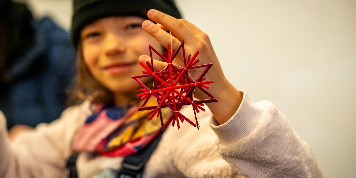 Ein Kind, das eine schwarze Mütze trägt, hält ein rotes geometrisches sternförmiges Ornament hoch und lächelt in die Kamera.