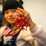 Ein Kind, das eine schwarze Mütze trägt, hält ein rotes geometrisches sternförmiges Ornament hoch und lächelt in die Kamera.