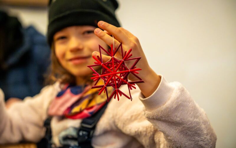 Ein Kind, das eine schwarze Mütze trägt, hält ein rotes geometrisches sternförmiges Ornament hoch und lächelt in die Kamera.