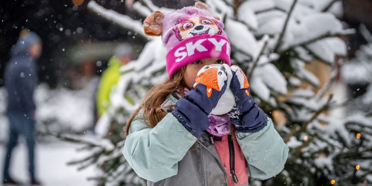 Ein Kind mit rosafarbener Mütze und Winterjacke trinkt vor einem verschneiten Baum aus einer Tasse; es schneit und die Menschen im Hintergrund sind unscharf.