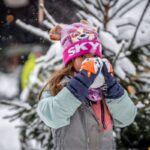 Ein Kind mit rosafarbener Mütze und Winterjacke trinkt vor einem verschneiten Baum aus einer Tasse; es schneit und die Menschen im Hintergrund sind unscharf.