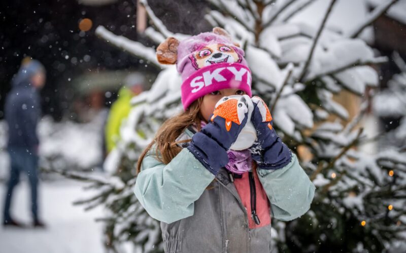 Ein Kind mit rosafarbener Mütze und Winterjacke trinkt vor einem verschneiten Baum aus einer Tasse; es schneit und die Menschen im Hintergrund sind unscharf.