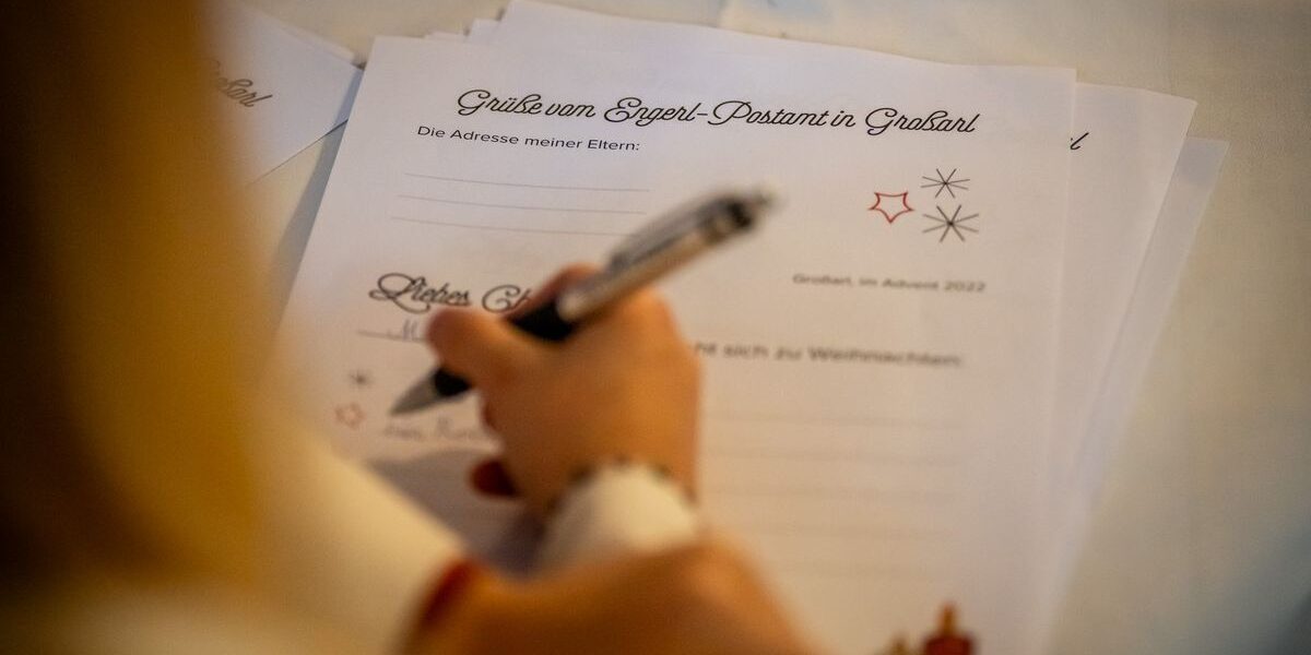 Eine Person schreibt auf einer Briefvorlage mit deutschem Text und der Überschrift "Grüße vom Engel-Postamt in Gradland", die auf einem Tisch liegt.
