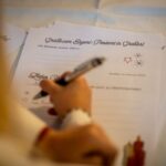 Eine Person schreibt auf einer Briefvorlage mit deutschem Text und der Überschrift "Grüße vom Engel-Postamt in Gradland", die auf einem Tisch liegt.