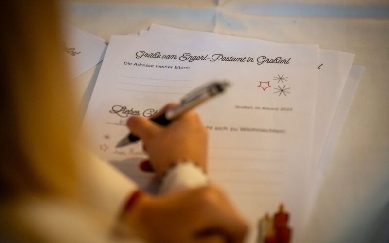 Eine Person schreibt auf einer Briefvorlage mit deutschem Text und der Überschrift "Grüße vom Engel-Postamt in Gradland", die auf einem Tisch liegt.