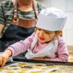 Ein kleines Kind mit einer Kochmütze verziert Kekse mit Streuseln auf einem Backblech, unterstützt von einem Erwachsenen in einer Schürze.