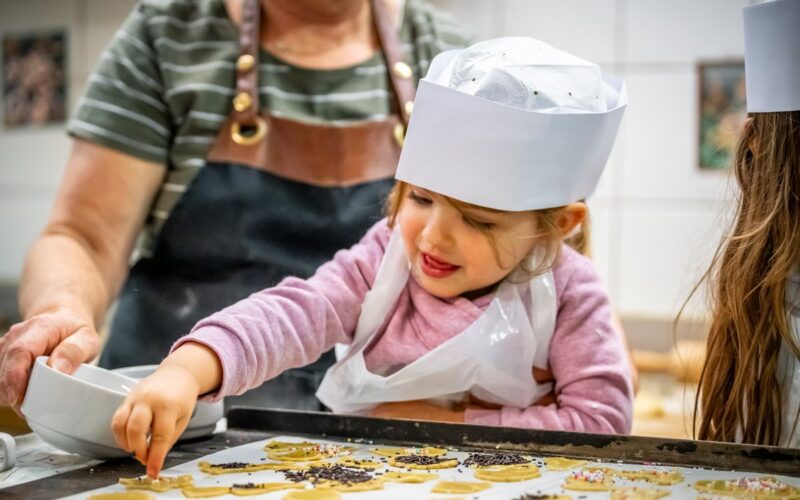 Ein kleines Kind mit einer Kochmütze verziert Kekse mit Streuseln auf einem Backblech, unterstützt von einem Erwachsenen in einer Schürze.
