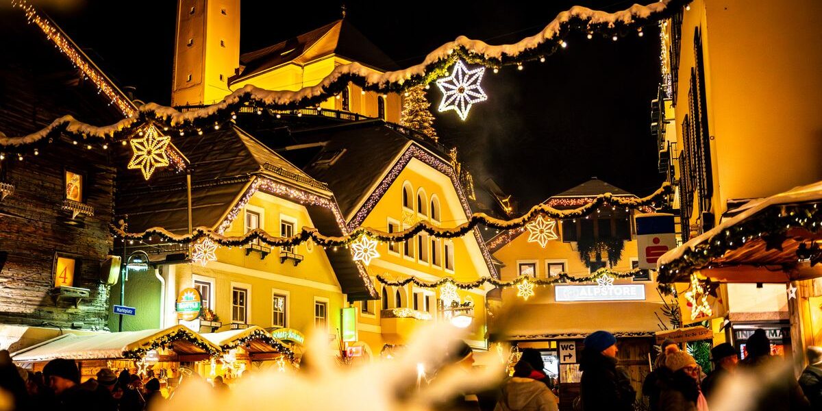 Eine festlich geschmückte Straße mit Lichtern und Sternenornamenten bei Nacht, mit Menschen und beleuchteten Gebäuden im Hintergrund.