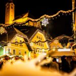 Eine festlich geschmückte Straße mit Lichtern und Sternenornamenten bei Nacht, mit Menschen und beleuchteten Gebäuden im Hintergrund.