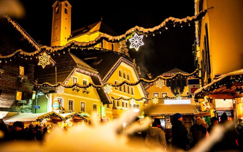 Eine festlich geschmückte Straße mit Lichtern und Sternenornamenten bei Nacht, mit Menschen und beleuchteten Gebäuden im Hintergrund.