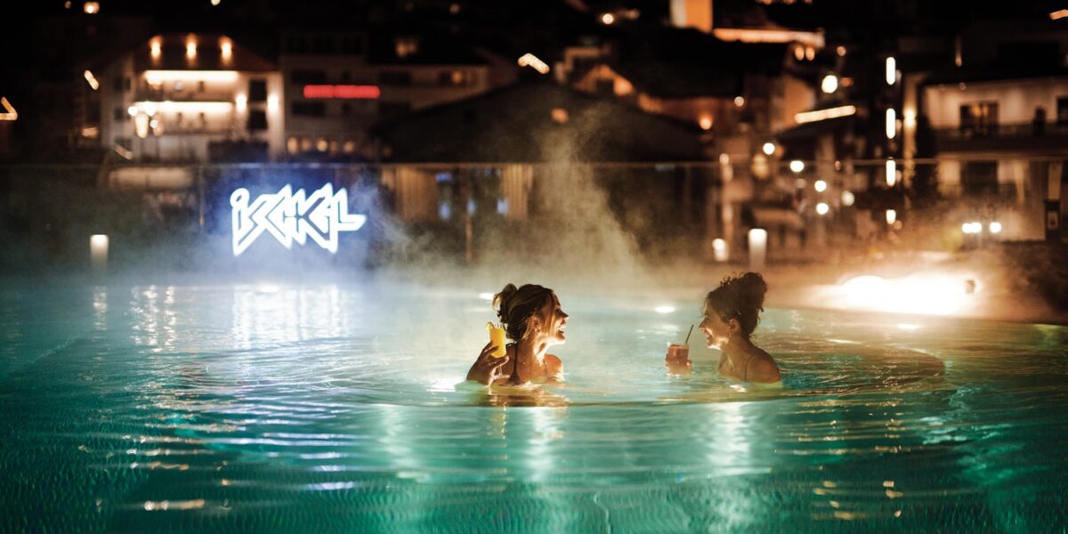 Zwei Personen befinden sich nachts in einem dampfenden Freibad, halten Getränke in der Hand und stehen sich gegenüber, während im Hintergrund Gebäude und Leuchtreklamen zu sehen sind.