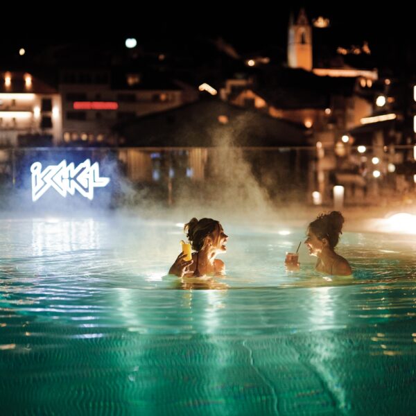 Zwei Personen befinden sich nachts in einem dampfenden Freibad, halten Getränke in der Hand und stehen sich gegenüber, während im Hintergrund Gebäude und Leuchtreklamen zu sehen sind.
