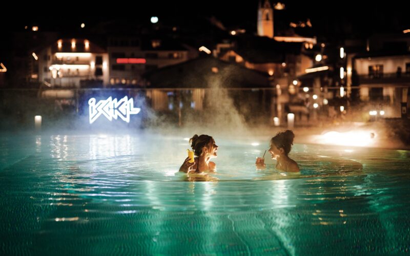 Zwei Personen befinden sich nachts in einem dampfenden Freibad, halten Getränke in der Hand und stehen sich gegenüber, während im Hintergrund Gebäude und Leuchtreklamen zu sehen sind.