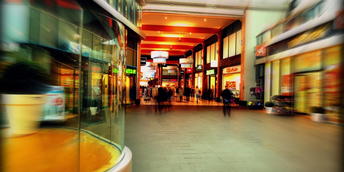 Ein hell erleuchtetes Einkaufszentrum mit verschiedenen Schaufenstern, beleuchteten Schildern und verschwommenen Gestalten, die den Korridor entlanggehen.