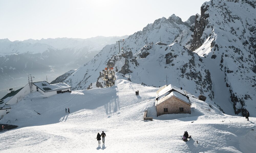 Menschen wandern auf einem schneebedeckten Berg mit mehreren Gebäuden und felsigen Gipfeln im Hintergrund unter einem hellen, klaren Himmel.