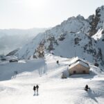 Menschen wandern auf einem schneebedeckten Berg mit mehreren Gebäuden und felsigen Gipfeln im Hintergrund unter einem hellen, klaren Himmel.