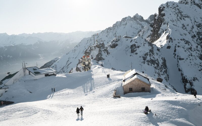 Menschen wandern auf einem schneebedeckten Berg mit mehreren Gebäuden und felsigen Gipfeln im Hintergrund unter einem hellen, klaren Himmel.