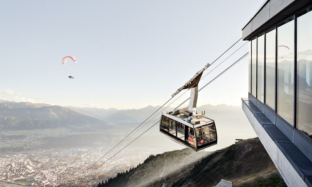 Eine Seilbahn startet von einer modernen Bergstation mit Blick auf eine Stadt und verschlungene Pfade, während ein Gleitschirm am Himmel über der Landschaft zu sehen ist.