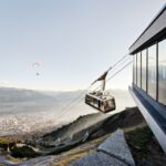 Eine Seilbahn startet von einer modernen Bergstation mit Blick auf eine Stadt und verschlungene Pfade, während ein Gleitschirm am Himmel über der Landschaft zu sehen ist.