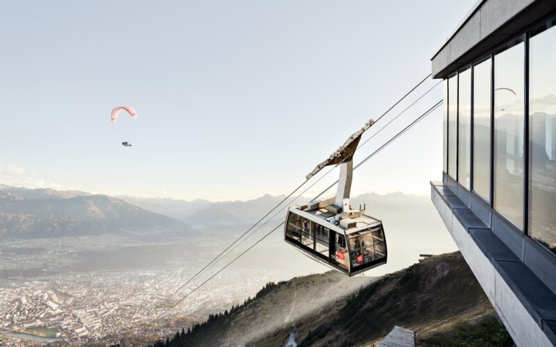 Eine Seilbahn startet von einer modernen Bergstation mit Blick auf eine Stadt und verschlungene Pfade, während ein Gleitschirm am Himmel über der Landschaft zu sehen ist.