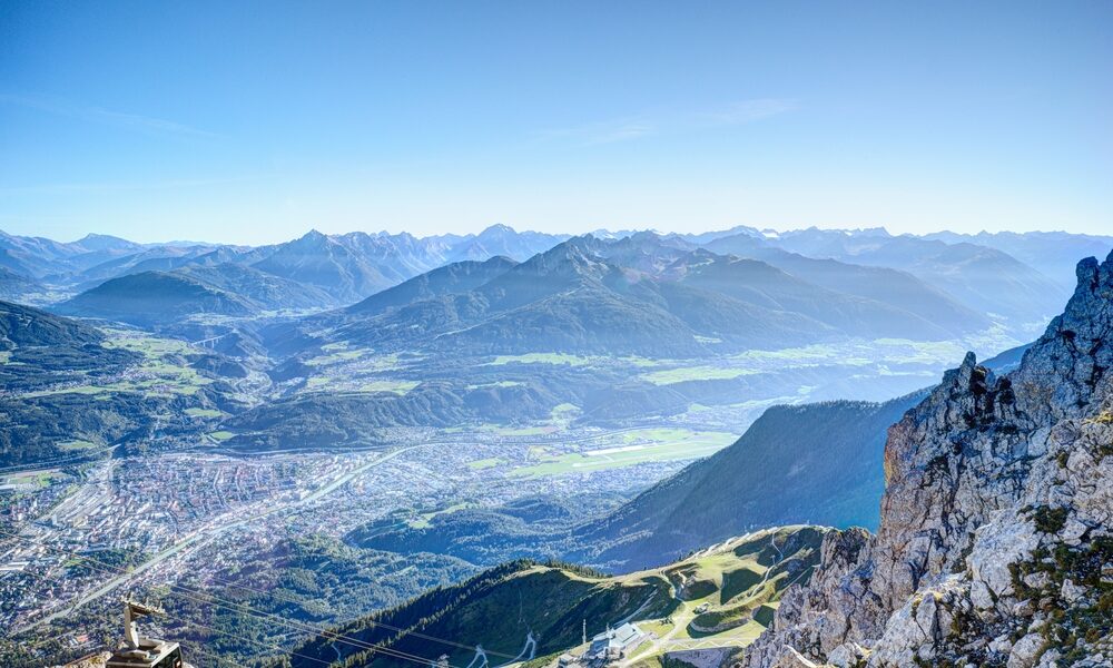 Blick von einem felsigen Berggipfel auf ein Tal mit einer Stadt, kurvenreichen Straßen und Bergketten in der Ferne unter einem klaren blauen Himmel.