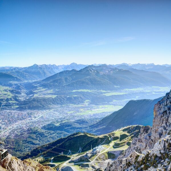 Blick von einem felsigen Berggipfel auf ein Tal mit einer Stadt, kurvenreichen Straßen und Bergketten in der Ferne unter einem klaren blauen Himmel.
