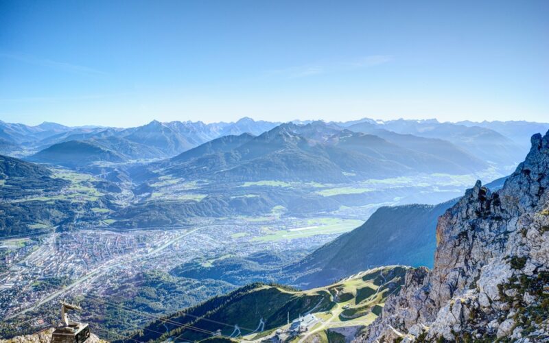 Blick von einem felsigen Berggipfel auf ein Tal mit einer Stadt, kurvenreichen Straßen und Bergketten in der Ferne unter einem klaren blauen Himmel.