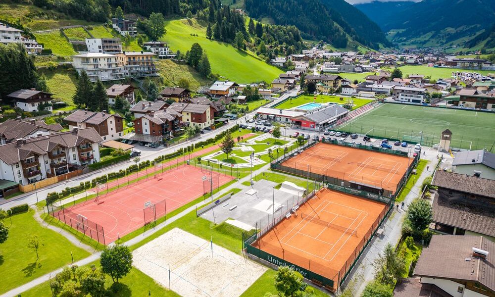 Luftaufnahme eines Sportkomplexes mit Sandtennisplätzen, einem Sandvolleyballplatz und einem Fußballfeld, umgeben von Häusern und grünen Hügeln in einem Bergdorf.