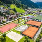 Luftaufnahme eines Sportkomplexes mit Sandtennisplätzen, einem Sandvolleyballplatz und einem Fußballfeld, umgeben von Häusern und grünen Hügeln in einem Bergdorf.
