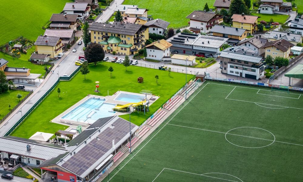 Luftaufnahme eines Fußballplatzes, eines Freibads mit Rutschen und nahe gelegenen Wohngebäuden in einer grünen, ländlichen Gegend.