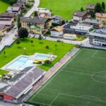 Luftaufnahme eines Fußballplatzes, eines Freibads mit Rutschen und nahe gelegenen Wohngebäuden in einer grünen, ländlichen Gegend.