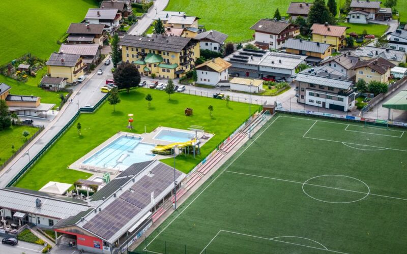 Luftaufnahme eines Fußballplatzes, eines Freibads mit Rutschen und nahe gelegenen Wohngebäuden in einer grünen, ländlichen Gegend.