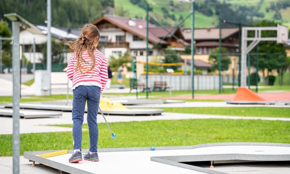 Ein junges Mädchen mit langen Haaren, das ein rot-weiß gestreiftes Hemd und Jeans trägt, spielt an einem sonnigen Tag im Freien in der Nähe von Gebäuden und grünen Hügeln Minigolf.
