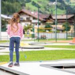Ein junges Mädchen mit langen Haaren, das ein rot-weiß gestreiftes Hemd und Jeans trägt, spielt an einem sonnigen Tag im Freien in der Nähe von Gebäuden und grünen Hügeln Minigolf.