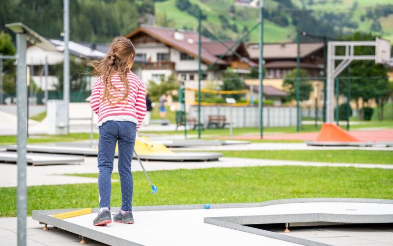 Ein junges Mädchen mit langen Haaren, das ein rot-weiß gestreiftes Hemd und Jeans trägt, spielt an einem sonnigen Tag im Freien in der Nähe von Gebäuden und grünen Hügeln Minigolf.