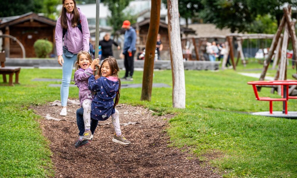 Zwei Kinder fahren gemeinsam auf einer Seilrutsche auf einem Spielplatz, während ein Erwachsener in der Nähe zusieht. Im Hintergrund sind andere Personen und Spielgeräte zu sehen.