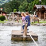 Zwei Mädchen stehen auf einem Holzfloß, das von Seilen über einen flachen Teich gezogen wird, in einer rustikalen Umgebung mit Hütten und Grünpflanzen im Hintergrund.