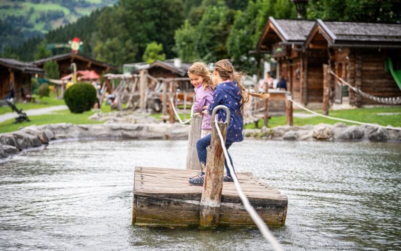 Zwei Kinder stehen auf einem Holzfloß und ziehen sich mit Hilfe von Seilen über einen flachen Teich, im Hintergrund sind Holzhütten und Grünflächen zu sehen.