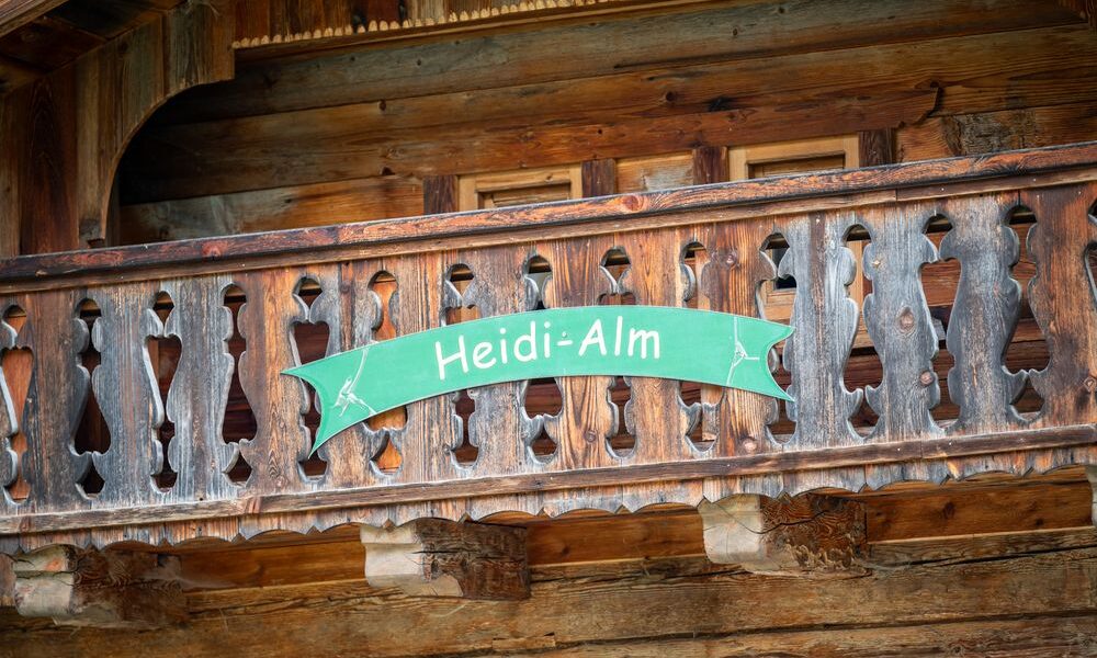Auf einem Holzbalkon mit geschnitztem Geländer steht ein grünes Schild mit der Aufschrift "Heidi-Alm".
