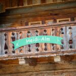 Auf einem Holzbalkon mit geschnitztem Geländer steht ein grünes Schild mit der Aufschrift "Heidi-Alm".