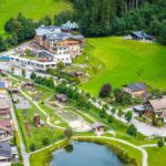 Luftaufnahme eines kleinen Dorfes mit Häusern, einem Teich, Spielplätzen, grünen Feldern und umliegenden Bäumen in der Nähe eines bewaldeten Hügels.