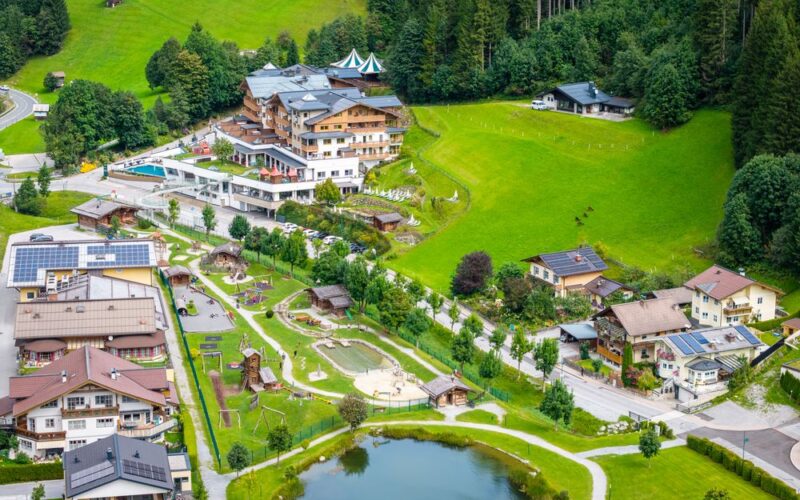 Luftaufnahme eines kleinen Dorfes mit Häusern, einem Teich, Spielplätzen, grünen Feldern und umliegenden Bäumen in der Nähe eines bewaldeten Hügels.