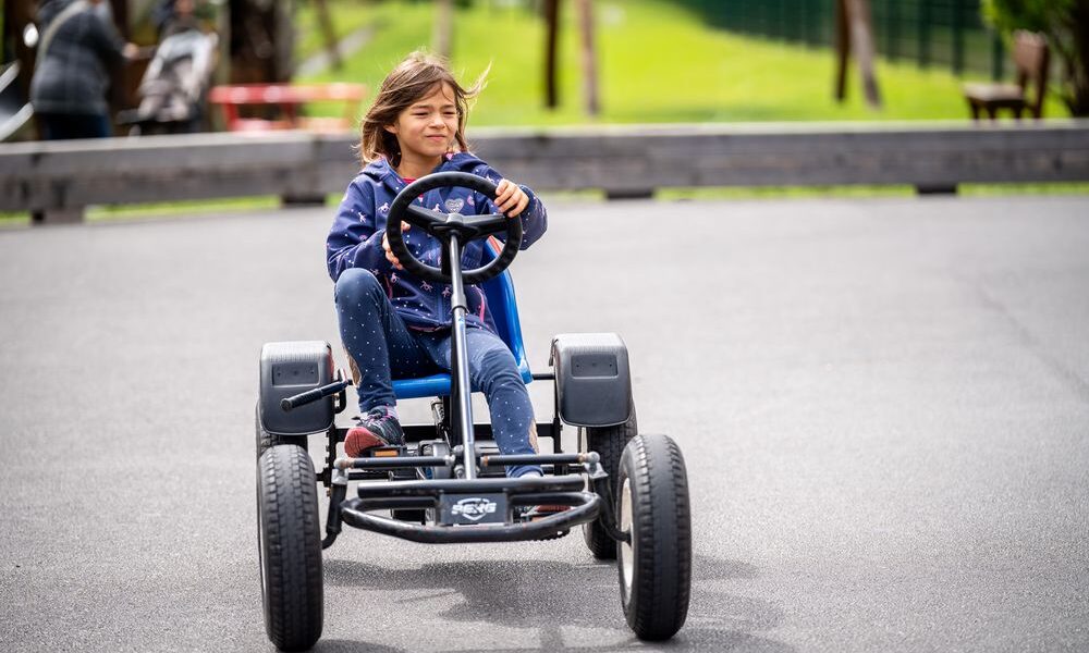 Ein Kind in blauem Kapuzenpulli und Jeans fährt mit einem Tret-Gokart auf einer gepflasterten Strecke im Freien, mit Grünflächen und Menschen im Hintergrund.