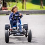 Ein Kind in blauem Kapuzenpulli und Jeans fährt mit einem Tret-Gokart auf einer gepflasterten Strecke im Freien, mit Grünflächen und Menschen im Hintergrund.