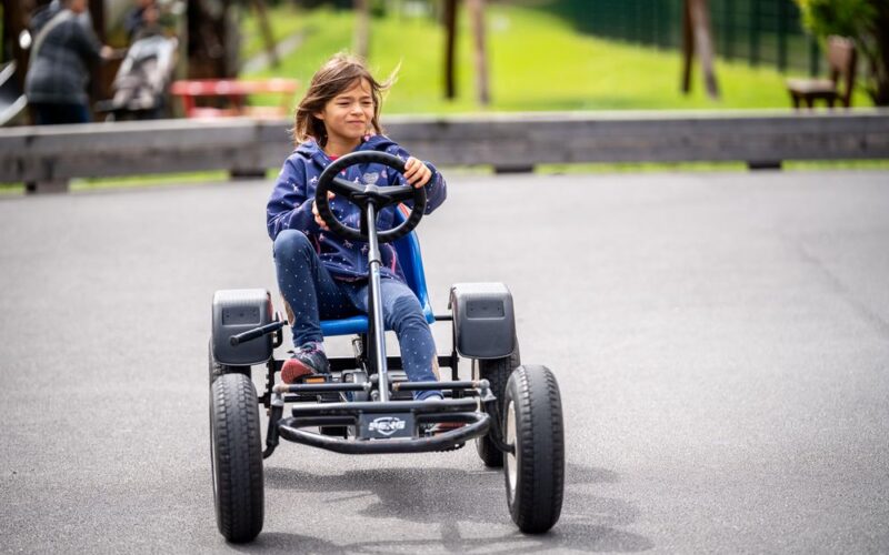 Ein Kind in blauem Kapuzenpulli und Jeans fährt mit einem Tret-Gokart auf einer gepflasterten Strecke im Freien, mit Grünflächen und Menschen im Hintergrund.