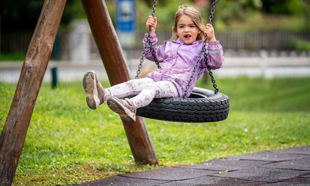 Ein junges Mädchen in einer lila Jacke sitzt auf einer Reifenschaukel auf einem Spielplatz, lächelt leicht und streckt die Beine aus, umgeben von Gras und Spielgeräten.