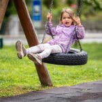 Ein junges Mädchen in einer lila Jacke sitzt auf einer Reifenschaukel auf einem Spielplatz, lächelt leicht und streckt die Beine aus, umgeben von Gras und Spielgeräten.
