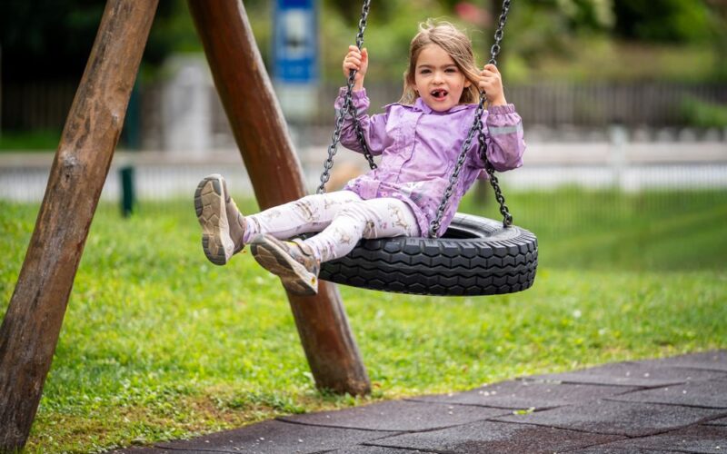 Ein junges Mädchen in einer lila Jacke sitzt auf einer Reifenschaukel auf einem Spielplatz, lächelt leicht und streckt die Beine aus, umgeben von Gras und Spielgeräten.